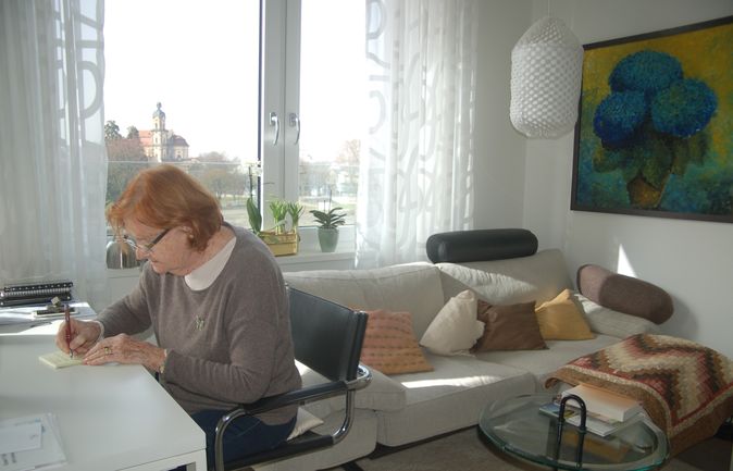 Ältere Frau schreibt am Schreibtisch vor Fenster mit Sofa im Hintergrund