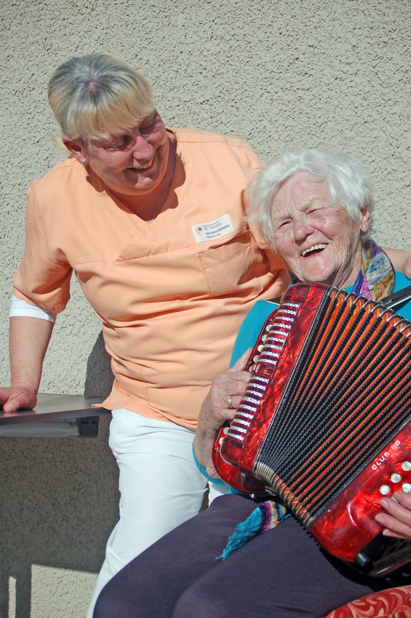 Betreuerin lacht mit Seniorin in Tagesbetreuung beim Akkordeonspielen