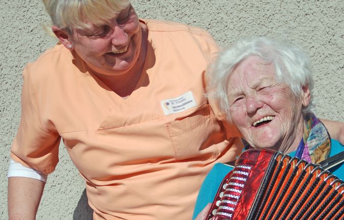 Betreuerin lacht mit Seniorin in Tagesbetreuung beim Akkordeonspielen