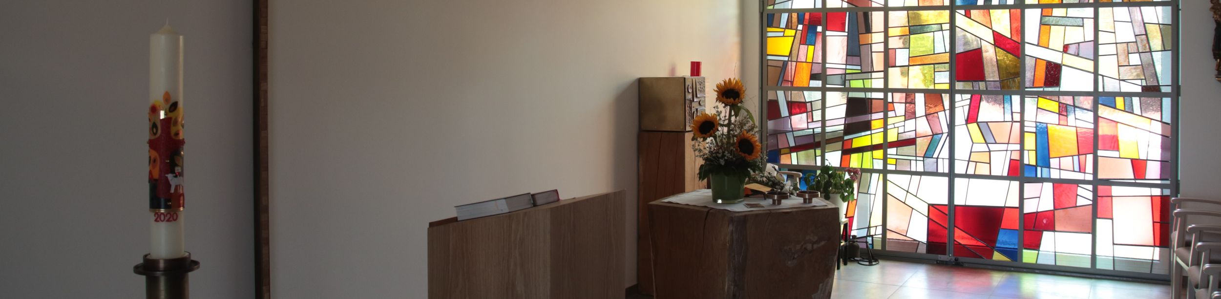 Innenraum einer Kapelle mit großem Holzkreuz, Altar, Sonnenblumen und buntem Glasfenster